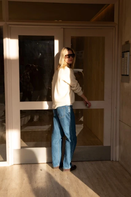 Adult woman with blonde hair, wearing a casual sweater and jeans, exits a building through glass double doors with sunlight hitting the frame.