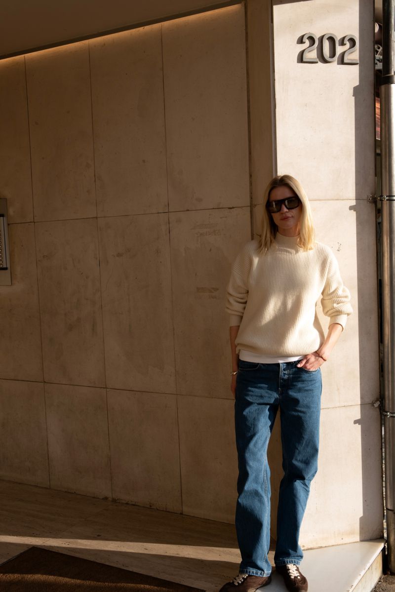 Woman in dark sunglasses wearing a cream sweater and blue jeans standing outdoors near a wall