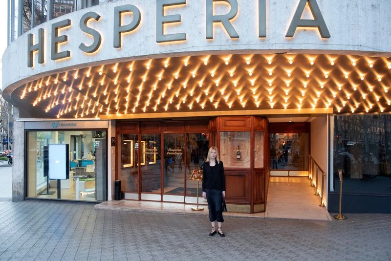 Full shot of a blonde woman standing in front of the illuminated Hesperia building in an urban setting, wearing a dark outfit.