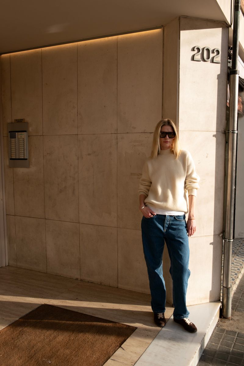 A woman in a cream sweater and jeans stands against a building with the number 202 visible. She has on sunglasses. The time of day is daytime.