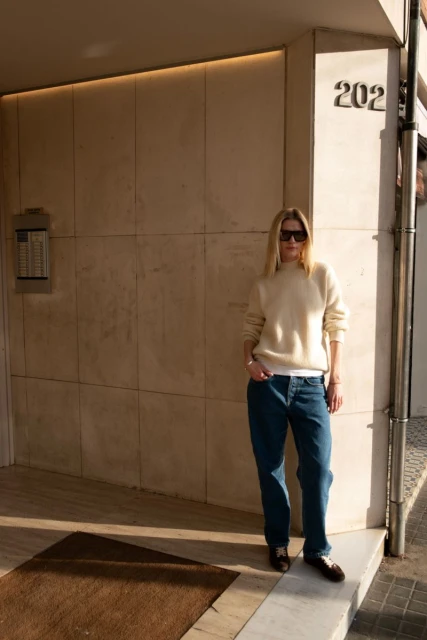 A woman in a cream sweater and jeans stands against a building with the number 202 visible. She has on sunglasses. The time of day is daytime.