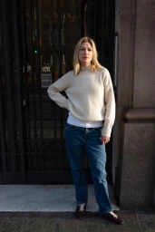 Woman in a cream knit sweater and blue jeans standing outside a building with large dark glass doors.