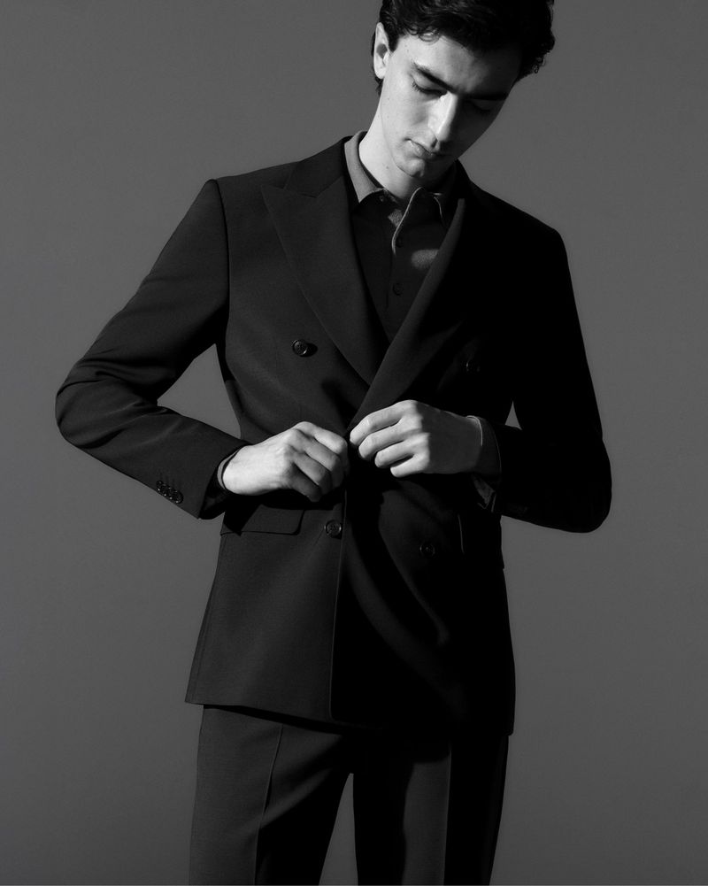Monochrome portrait of a young man in a dark double-breasted suit buttoning the jacket, posing against a dark grey backdrop in a photo studio.
