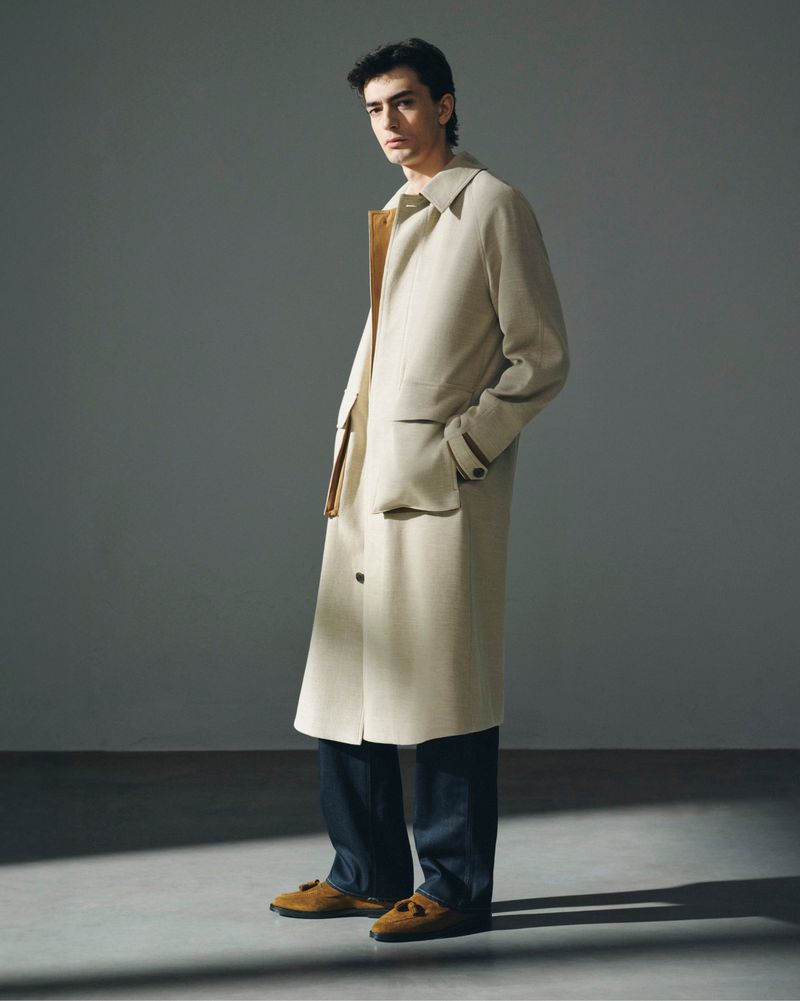 A fashionable adult male poses indoors wearing a light beige trench coat, dark blue pants, and brown loafers against a grey background.
