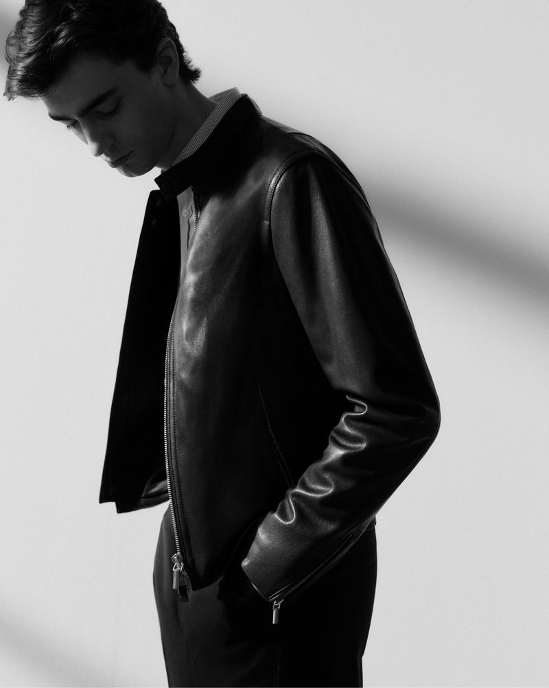 Black and white portrait of a young adult male wearing a dark leather jacket with hands in his pockets, minimalist fashion style.