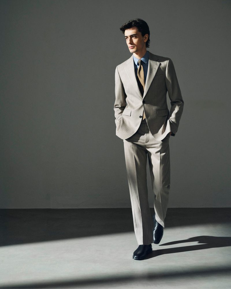 A stylish adult male in a grey suit walks toward the viewer in a brightly lit studio, creating a stark shadow and fashion-forward look.
