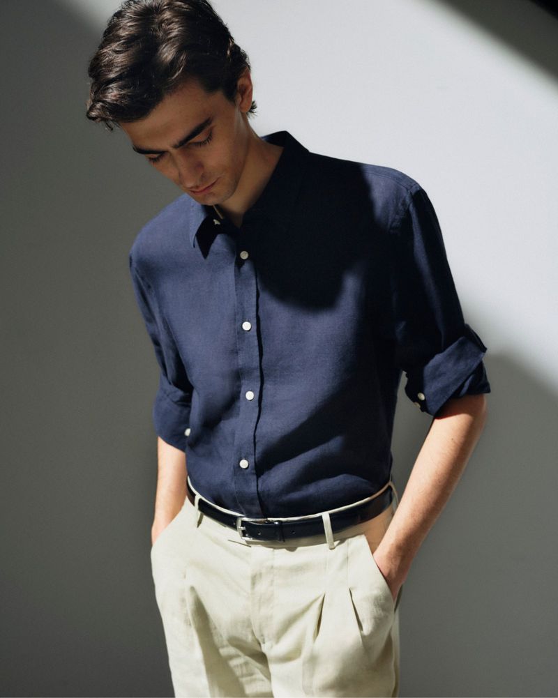 A male fashion model is posing in a blue linen button-down shirt and beige pleated trousers in an indoor setting with soft lighting.