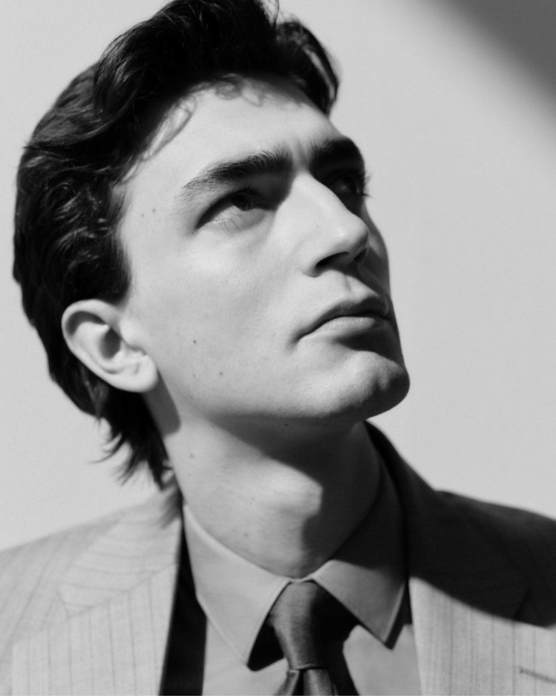 Portrait of a light-skinned adult male in a striped suit, collared shirt, and tie. He gazes upwards in a black-and-white studio shot.