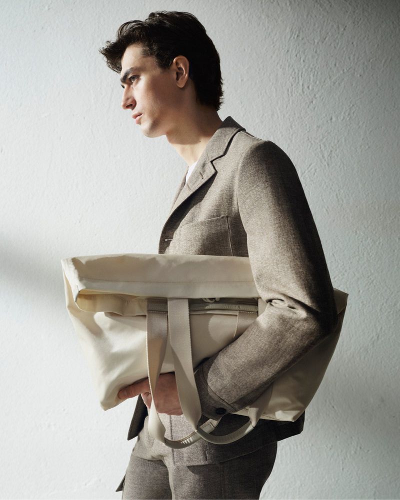 Adult man in suit jacket holds neutral tote bag indoors against a textured white wall in a fashion or lifestyle context.