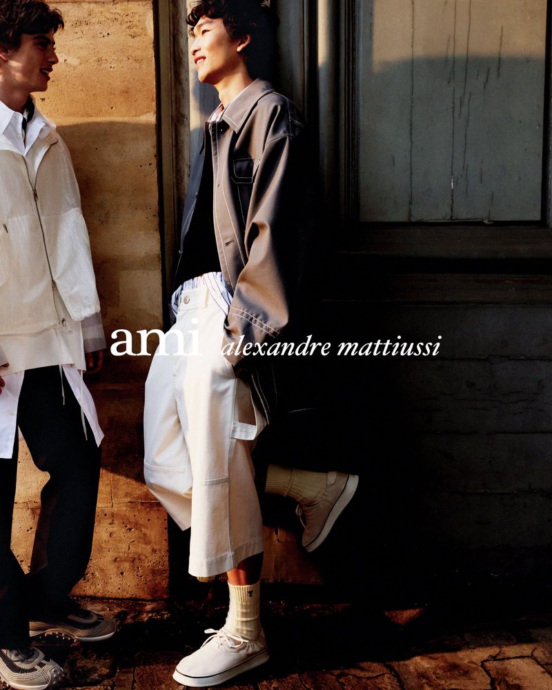 Two adult male models pose in neutral-toned casual fashion leaning against a weathered wall outside in natural light, editorial fashion.