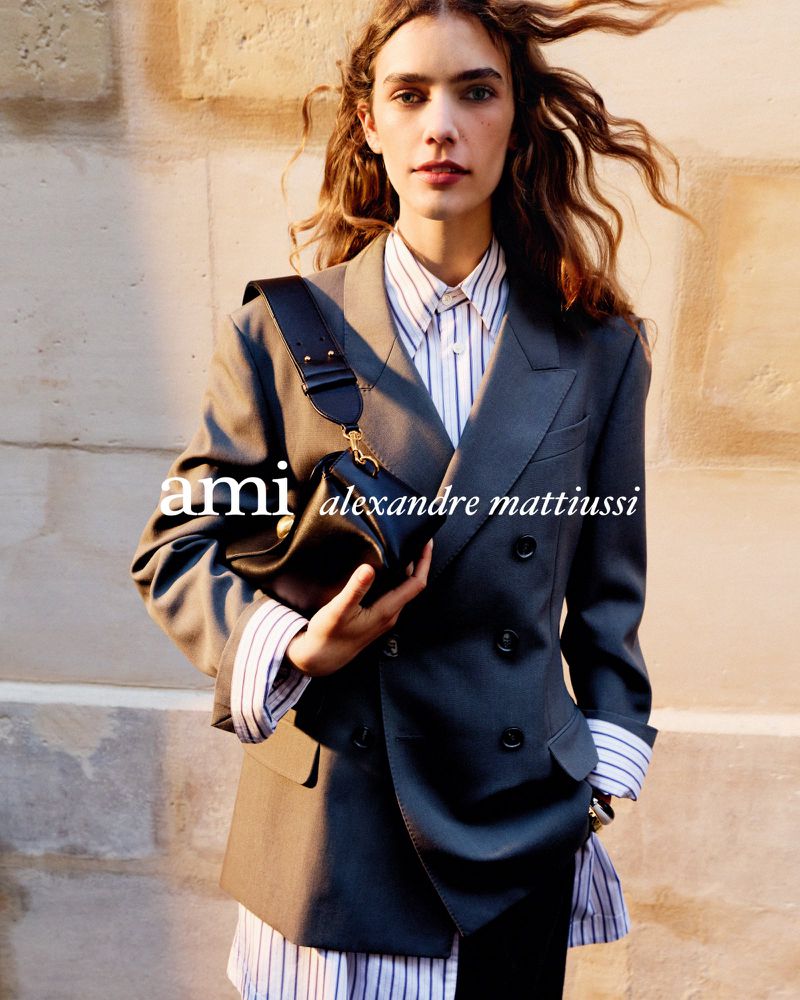 A fashionable young woman poses confidently wearing an oversized grey blazer, a striped shirt, and a crossbody bag against a textured wall.