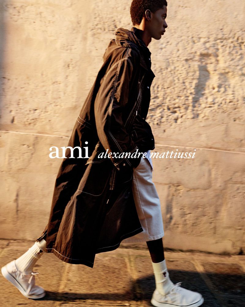 Adult male fashion model walks on a city street wearing a long dark hooded coat, white shorts, white socks, and white sneakers.