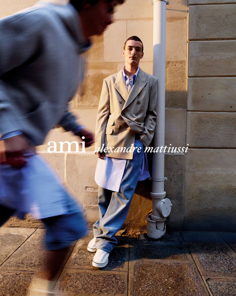 Fashion shot of a man in a beige blazer and jeans leaning against a wall, another man in motion is in the blurred background.