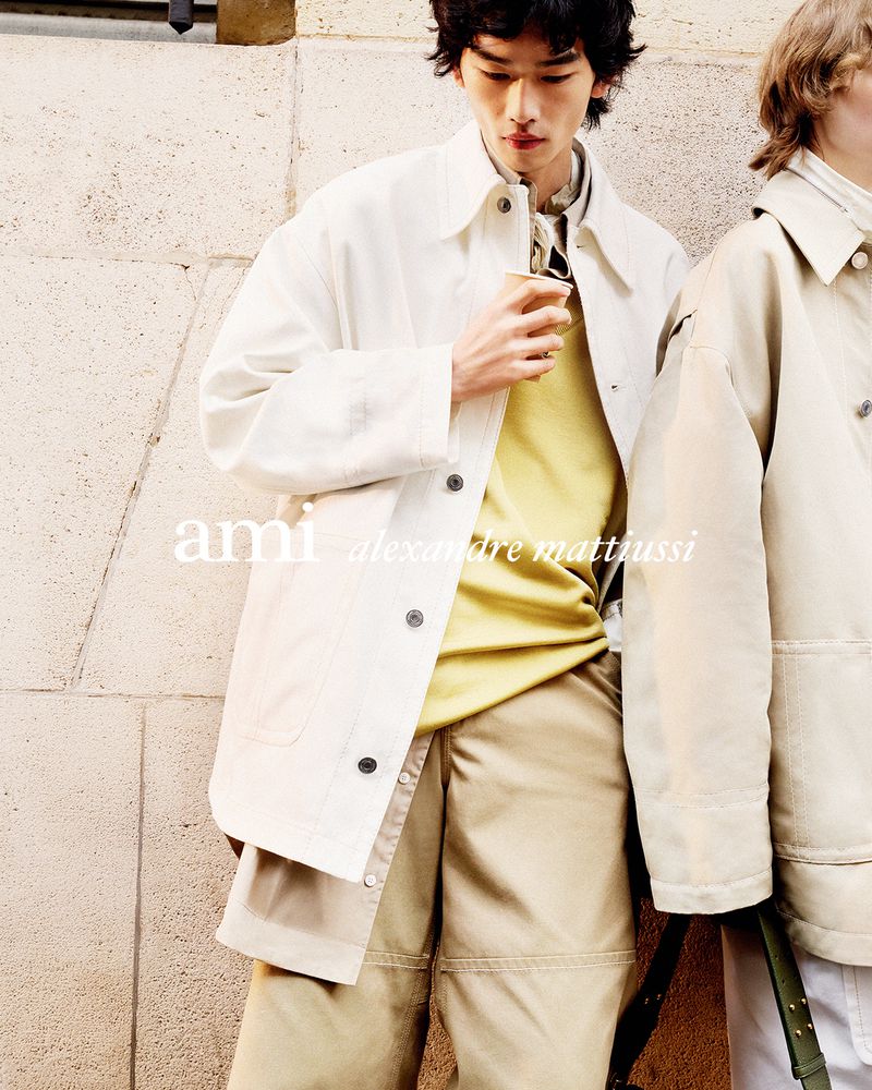 Two men in beige and white jackets stand against a textured wall, showcasing fashion apparel in a soft, natural outdoor light.
