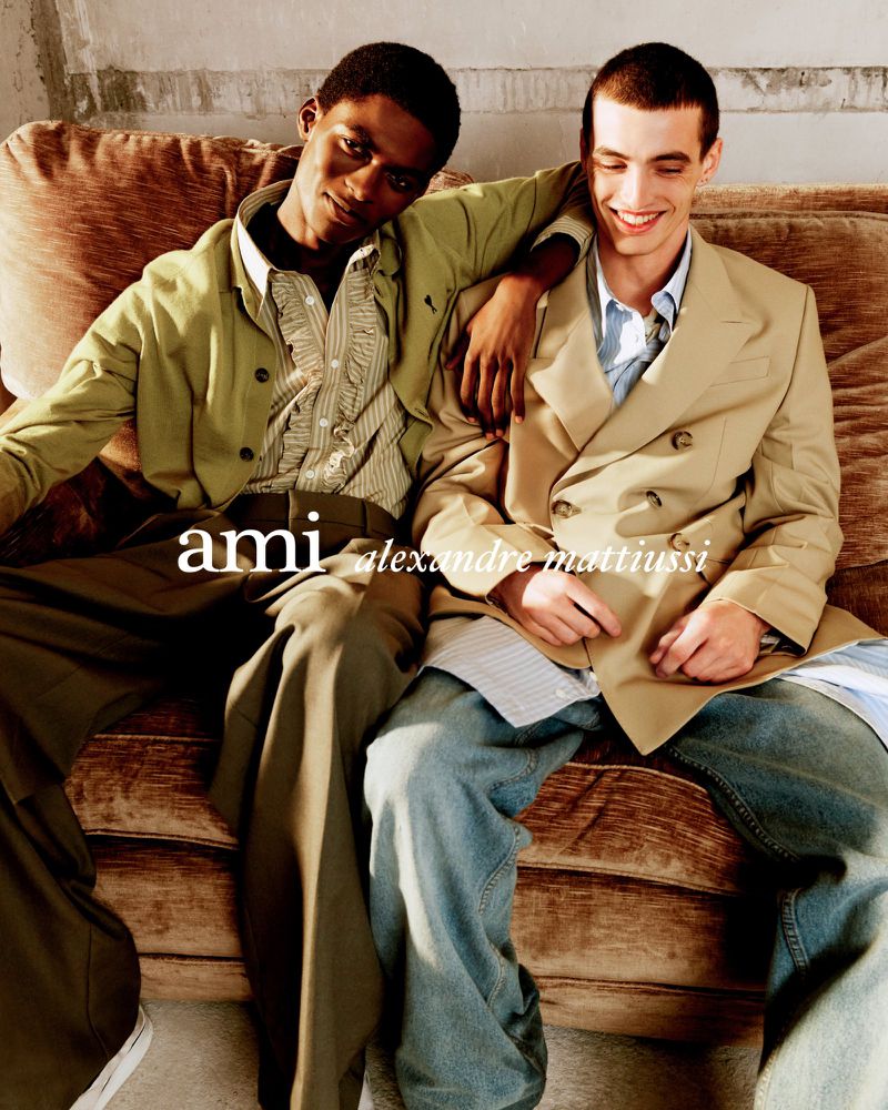 Two male fashion models pose on a brown couch wearing stylish coats and jeans in a studio shot for an ad, showcasing fashion.