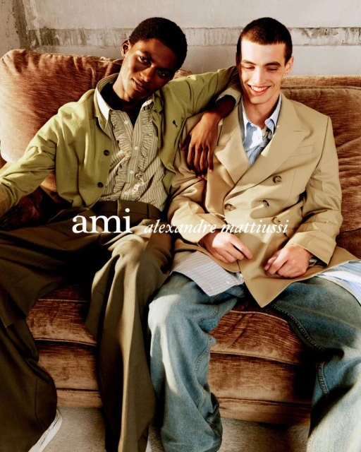 Two male fashion models pose on a brown couch wearing stylish coats and jeans in a studio shot for an ad, showcasing fashion.