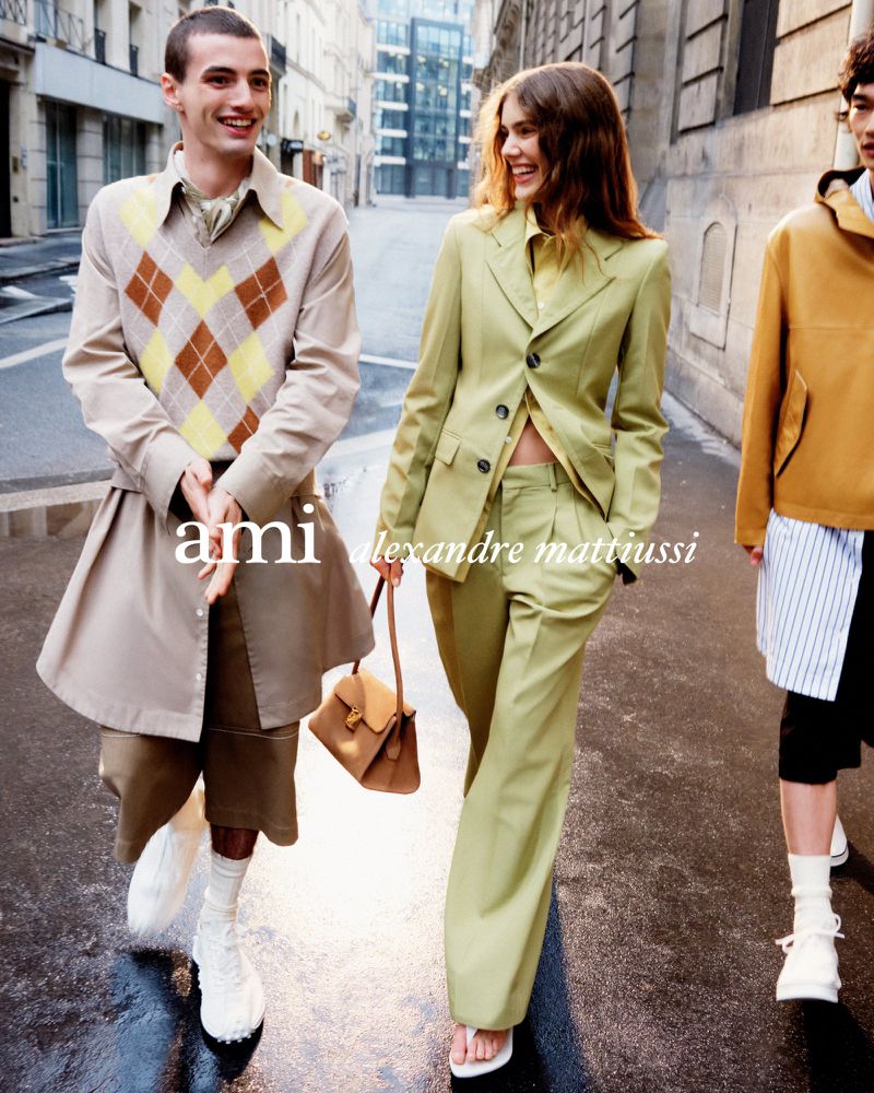 Fashion editorial featuring a stylish group of three young adults in designer clothing, walking on a wet city street with buildings.
