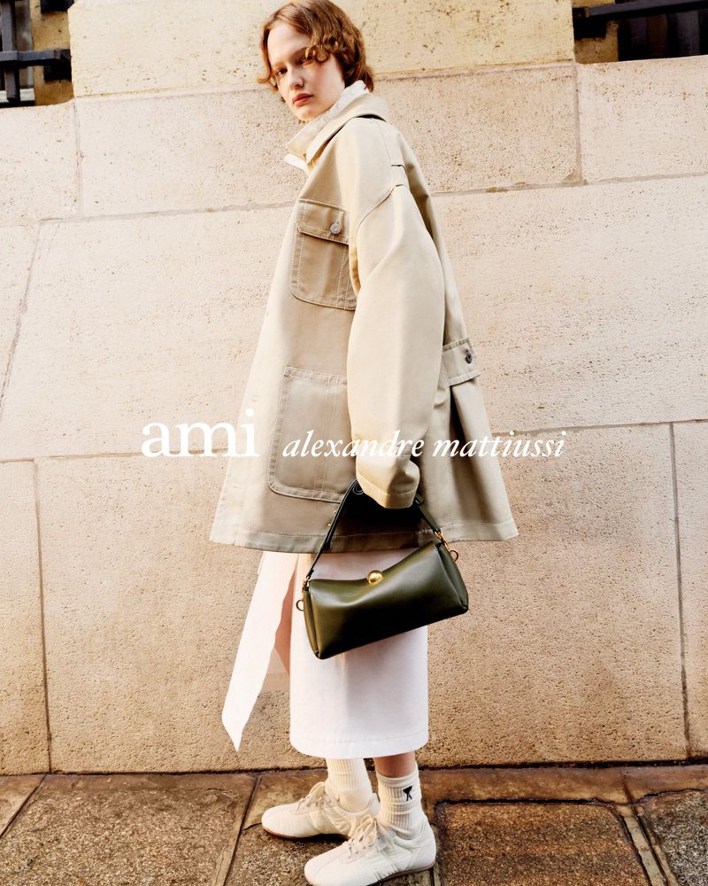Model wearing a cream jacket and holding a dark green handbag in front of a stone wall for the Ami S/S 2026 campaign.