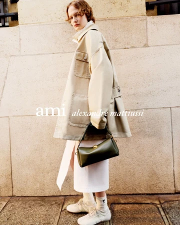 Model wearing a cream jacket and holding a dark green handbag in front of a stone wall for the Ami S/S 2026 campaign.