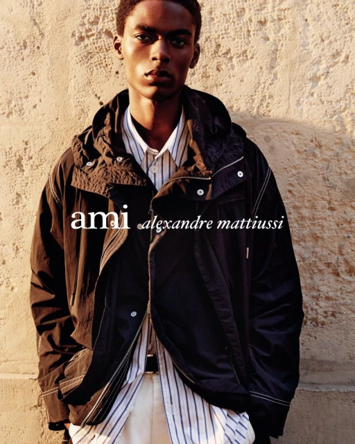 A young Black male model wears an open black jacket, striped shirt, and cream pants against a textured tan wall in an outdoor fashion shot.