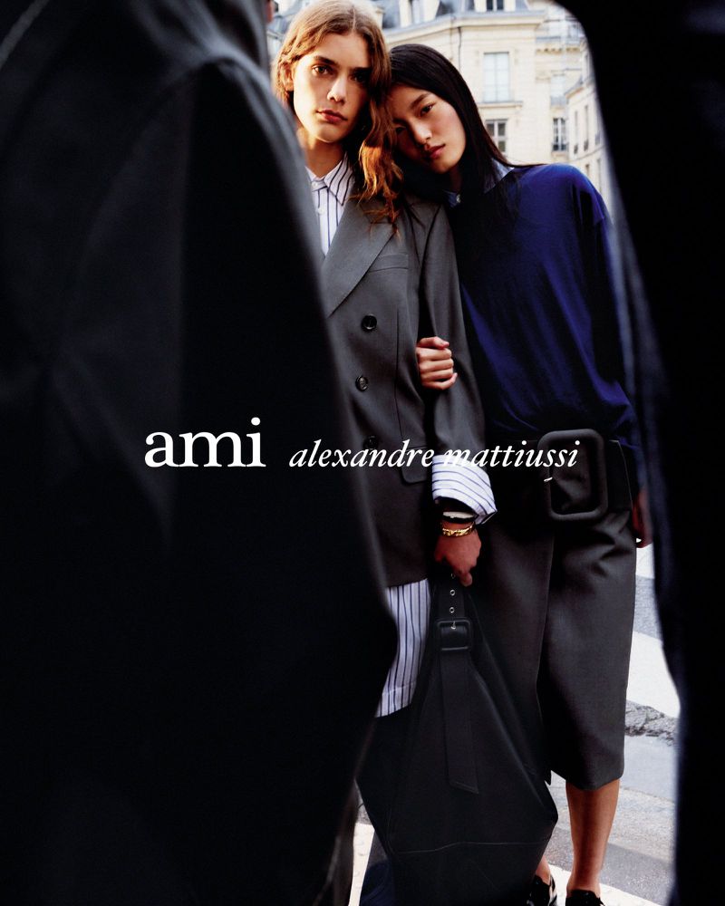 Two fashion models in Ami Alexandre Mattiussi clothing pose in an urban setting with visible buildings in the background.