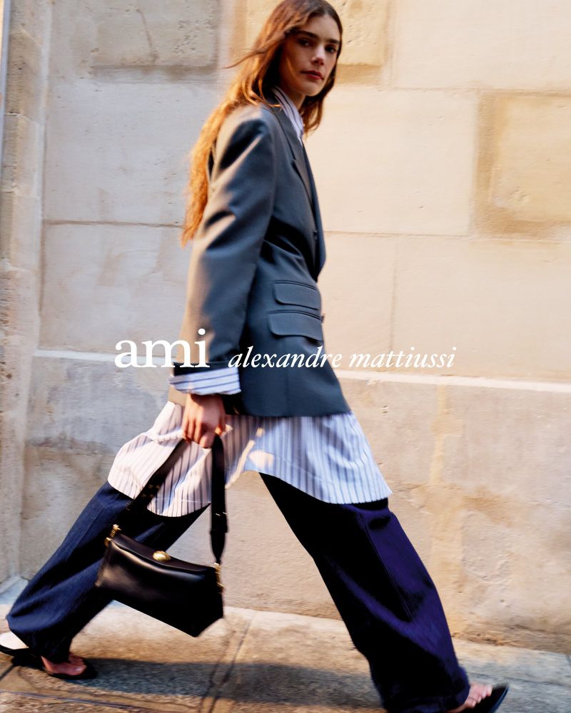 Model walking outdoors wearing an oversized grey blazer, striped shirt, and wide-leg trousers while carrying a black leather handbag.