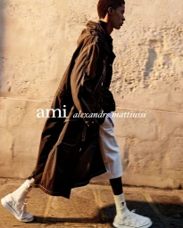 A Black man walking past a textured stone wall wearing a long dark hooded coat, light shorts, and white sneakers.
