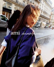 Model in a blue sweater carrying a black handbag walking on a Paris street for the Ami Spring/Summer 2026 campaign.