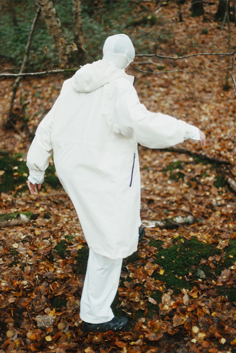 Person in a white hooded suit walks on mossy rocks amongst brown leaves in a sparse forest setting, viewed from behind.