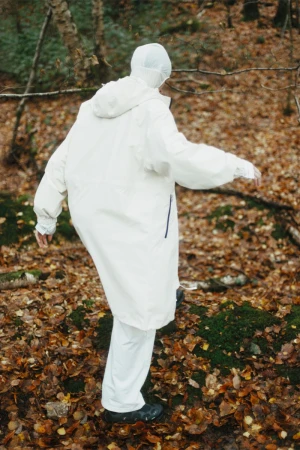 Person in a white hooded suit walks on mossy rocks amongst brown leaves in a sparse forest setting, viewed from behind.