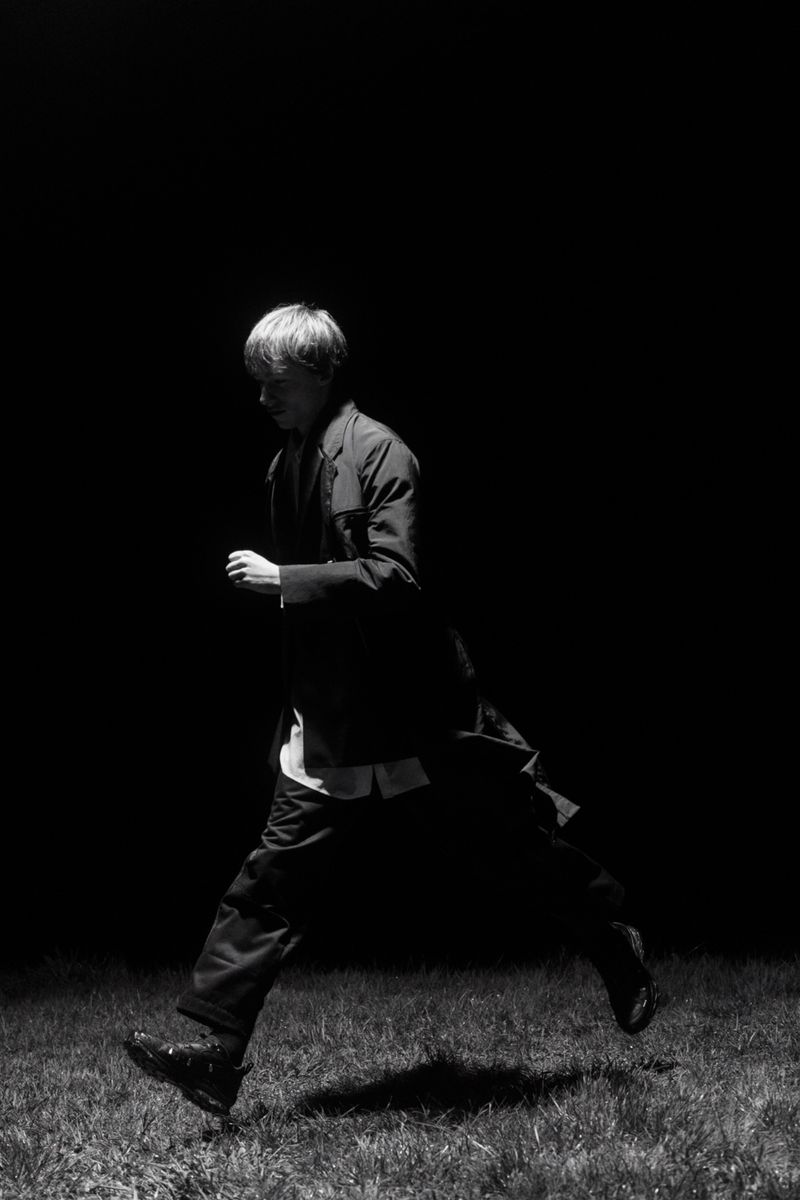 A person in a dark jacket and pants is captured mid-air running across a grassy field, creating a dynamic black-and-white image.