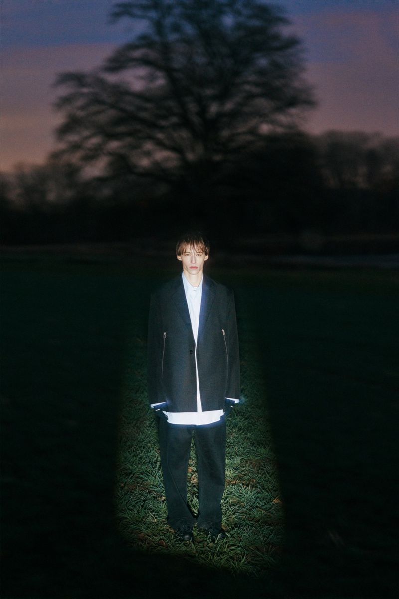 Full-shot of adult male standing in a field at dusk wearing a dark jacket and pants. A dark tree is in the background.