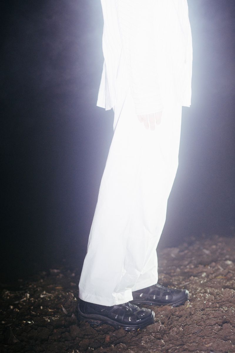 Person in white suit stands on dirt ground in a dark setting, bathed in bright light with shiny black shoes, studio shot, fashion style.