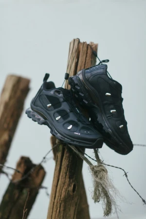 Two black athletic shoes are displayed on wooden posts wrapped with barbed wire, set against a neutral background in a product shot.