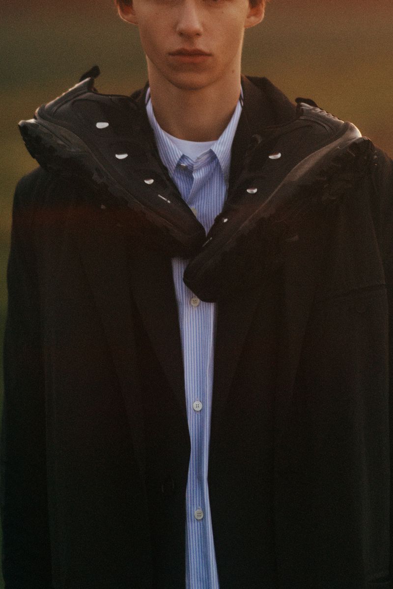 Fashion shot of a young adult man in a dark coat and striped shirt, with two dark sneakers draped over his shoulders, set outdoors.