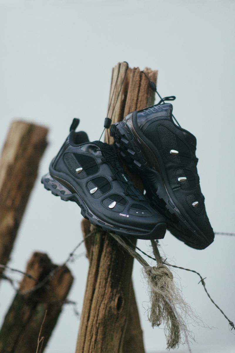 Pair of black OAMC x Salomon sneakers hanging by laces from a weathered wooden post outdoors.