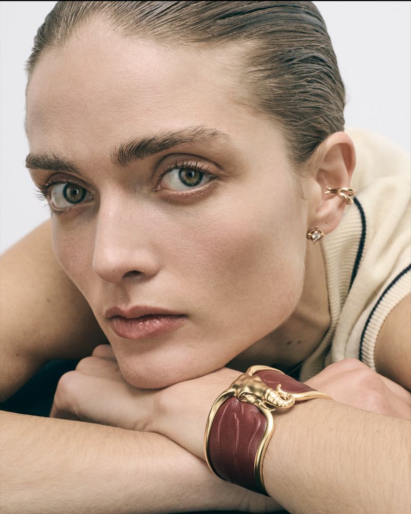 Close-up portrait of a woman wearing a burgundy and gold cuff bracelet with an elephant head, gold hoop earrings, and a collared shirt.