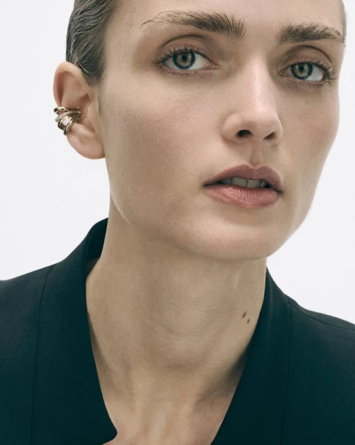 Close-up of a woman with green eyes wearing a gold ear cuff on her left ear and a black blazer against a plain white background.