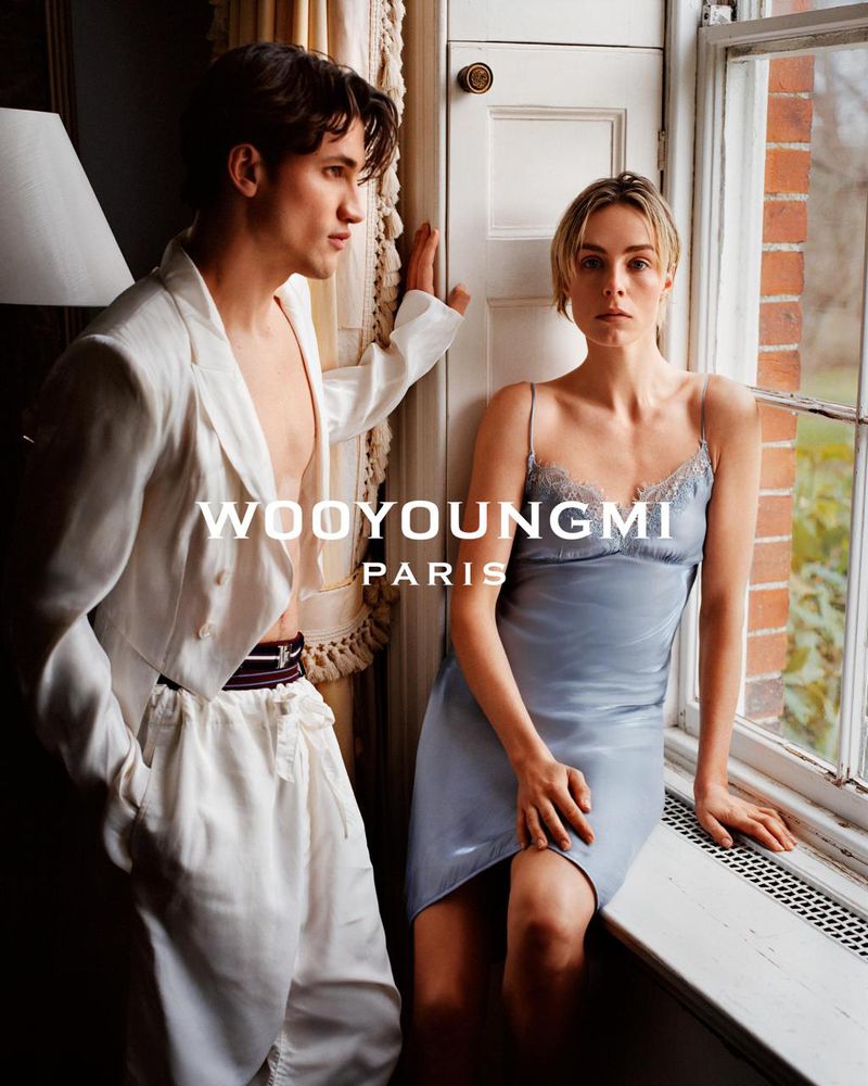 Two fashion models pose indoors near a window. The man wears a white blazer, and the woman wears a satin dress in this eye-level shot.