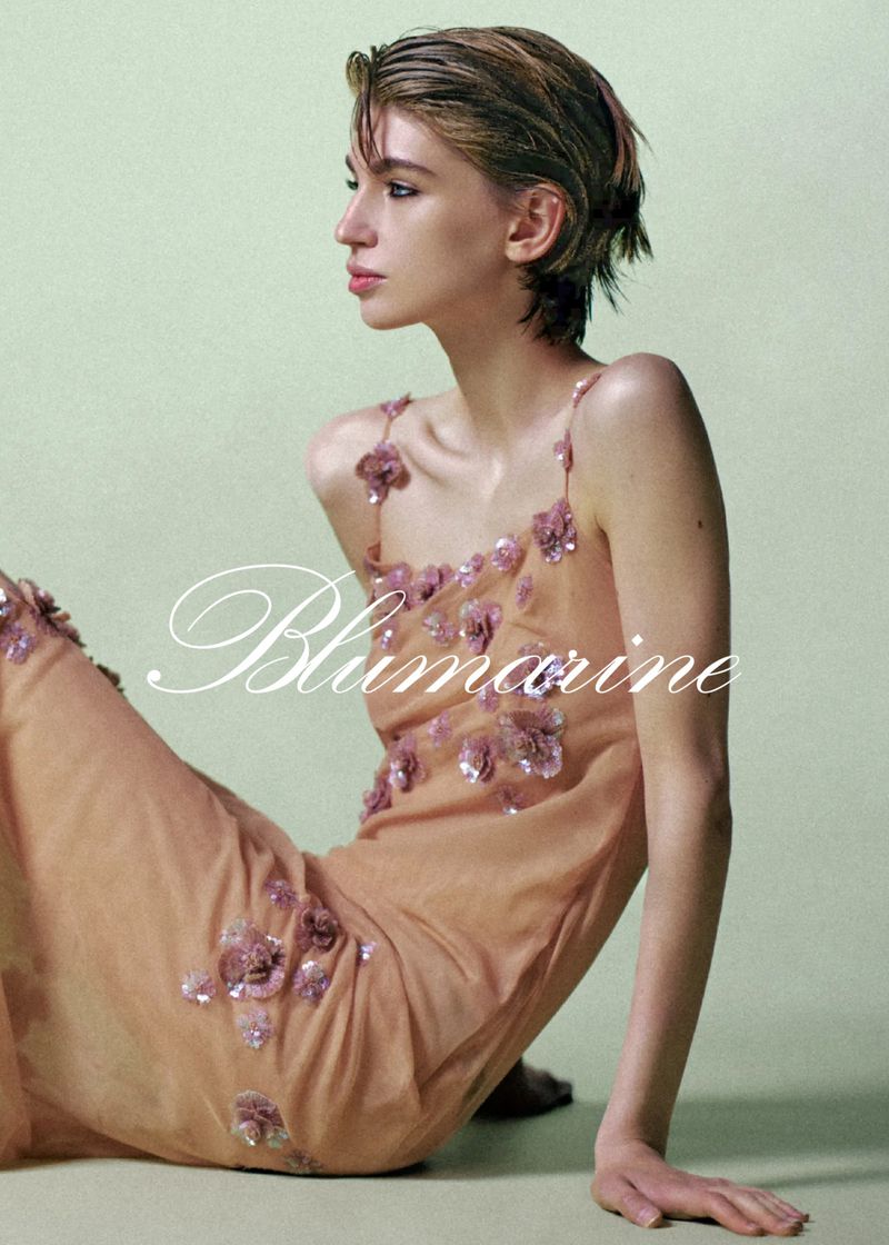 A young adult woman with short hair poses in a peach colored gown with floral accents against a light green background.