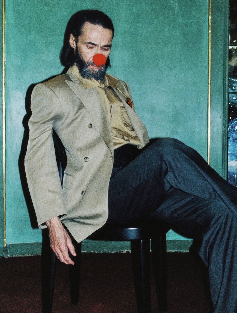 Adult male with a beard wearing a suit and a red clown nose, sitting on a stool in front of a green wall. The lighting is soft.