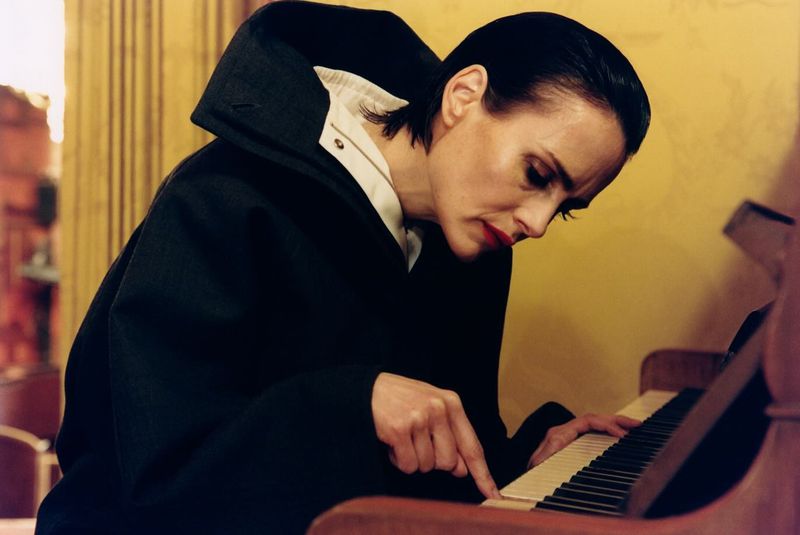 Adult female plays piano indoors in a black hooded jacket and white collared shirt, looking at keys in a close up eye level shot.