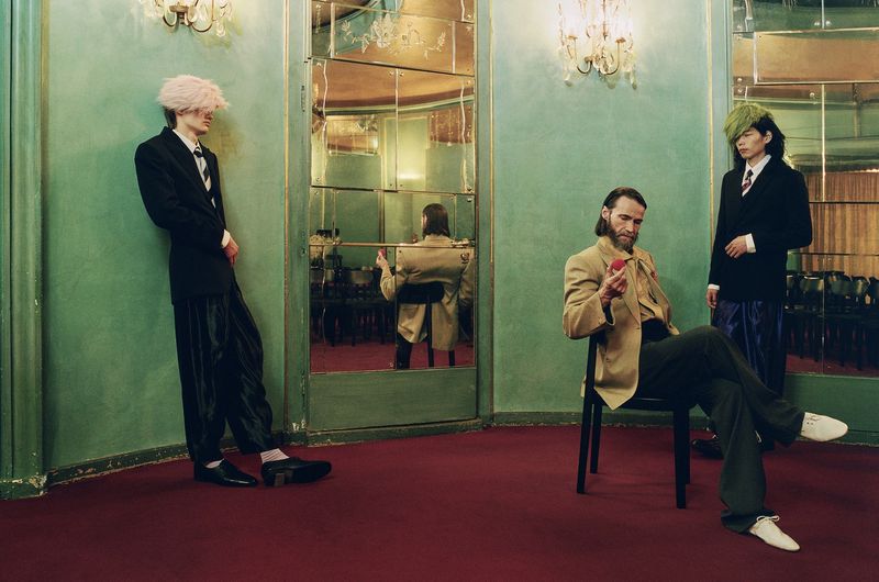 Three stylishly dressed adult men pose in a room with light green walls, red carpet, and vintage decor, illuminated by elegant chandeliers.
