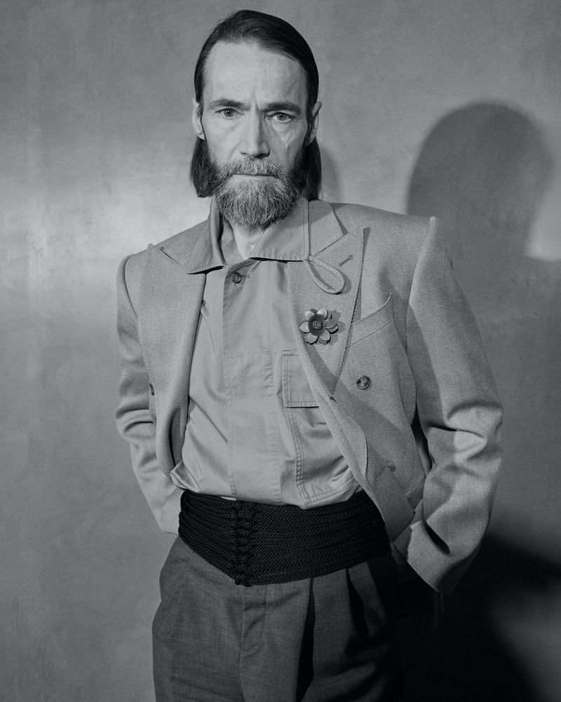 Black and white portrait of an adult male with a beard wearing a suit jacket, shirt, and decorative belt against a textured wall.
