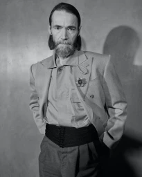 Portrait of an elderly man with a beard wearing a structured blazer and cummerbund against a plain wall.