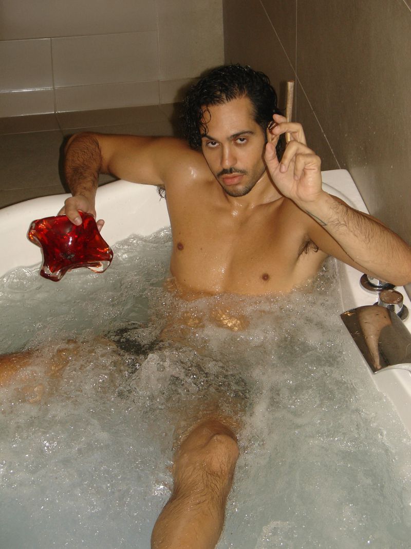 Adult male soaks in bubbly hot tub with a red glass bowl, showcasing relaxation and self-care in a minimalist tiled bathroom setting.