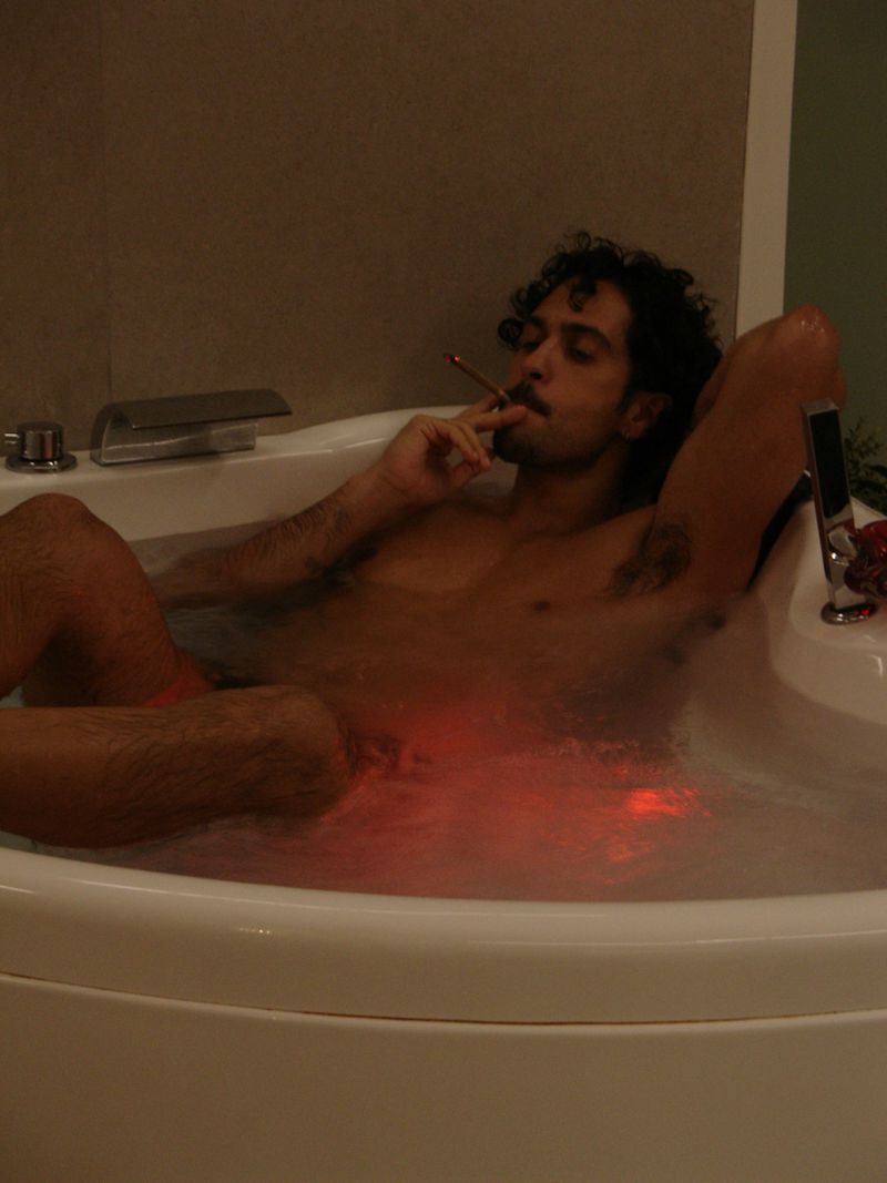 Man relaxes in a filled jacuzzi with a cigarette. The indoor setting has minimal decoration with neutral background wall colours.