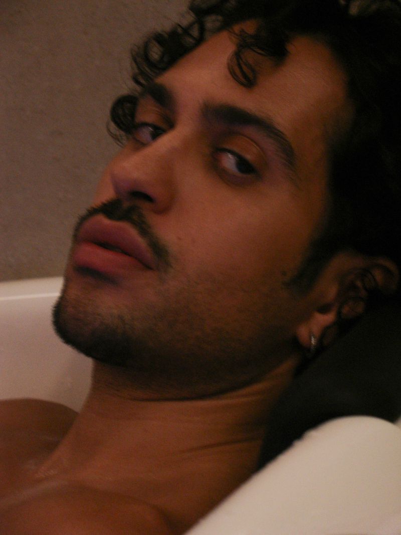 Close-up portrait of a reclining adult male with dark curly hair, mustache, and beard, fair skin tone, resting on a white surface indoors.