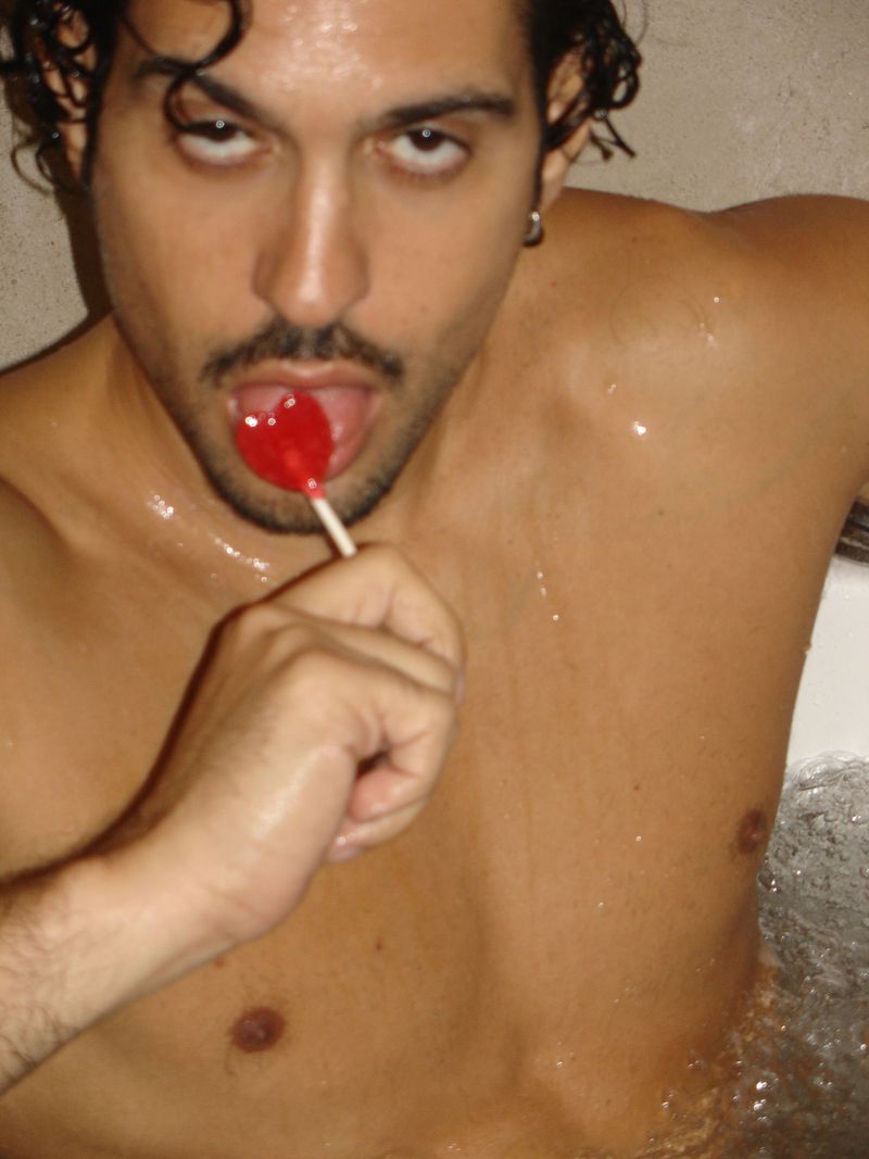 Adult male poses shirtless indoors in a bathtub, holding and licking a red lollipop; he has dark hair and fair skin.