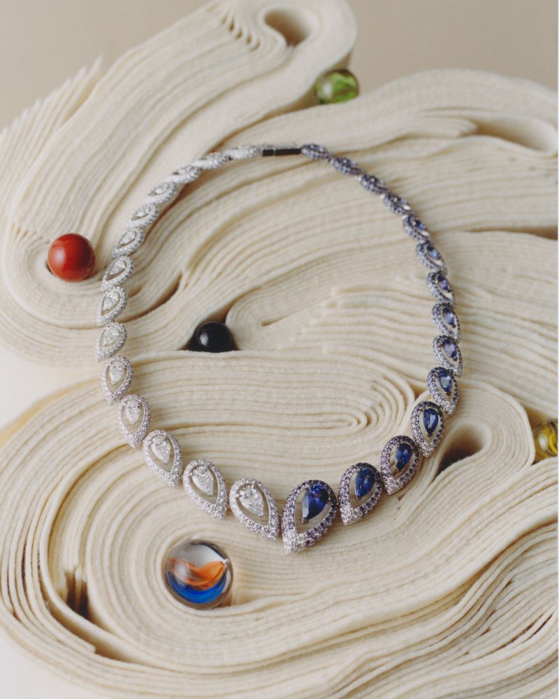 Elegant teardrop necklace featuring diamonds and sapphires, displayed on a textured surface with decorative glass marbles, jewellery shot.